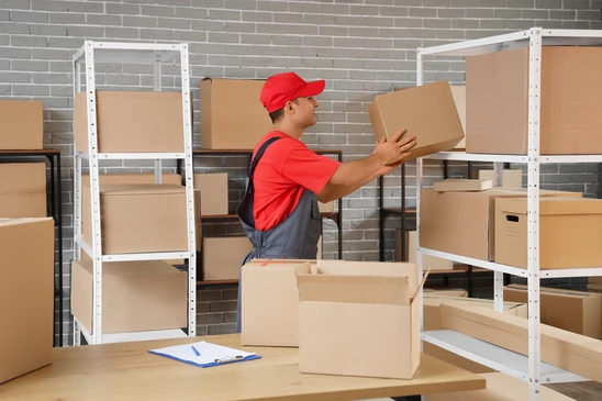 Livreur en uniforme tenant un colis cartonné dans un entrepôt logistique avec pile de cartons d'expédition